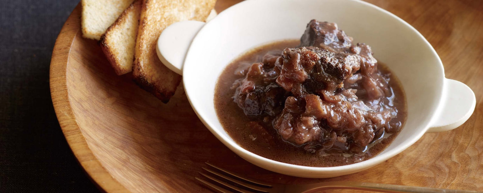 Rich beef stew in a dark sauce, served in a white bowl with toasted bread slices on the side.