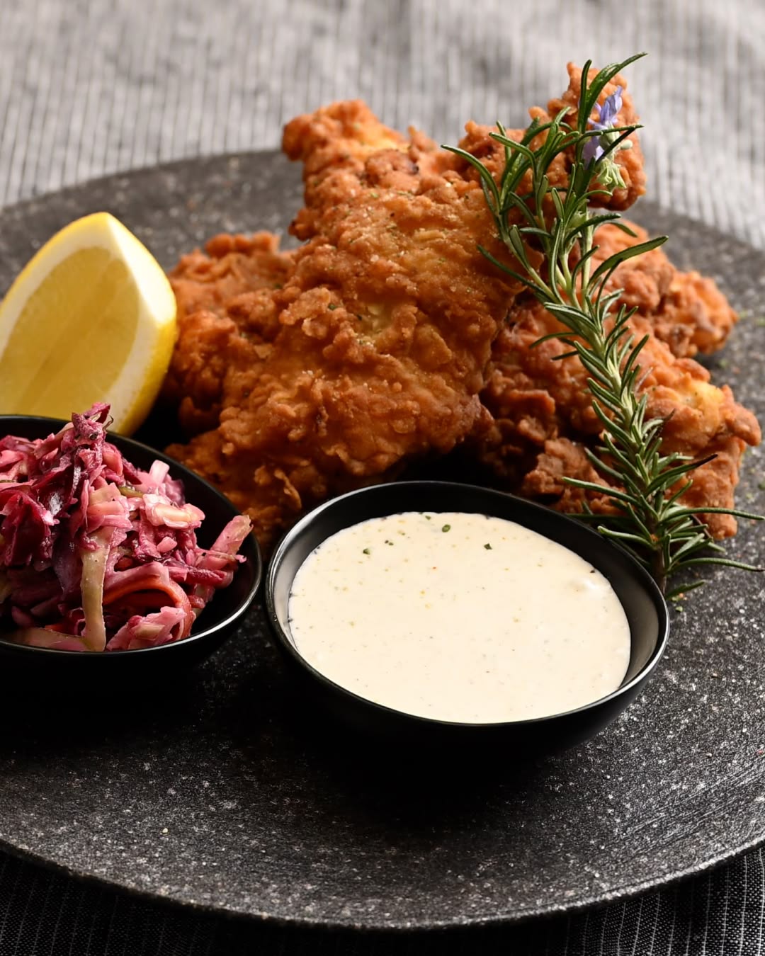 Crispy fried chicken pieces served with lemon wedge, rosemary garnish, creamy dipping sauce, and a side of purple cabbage slaw.