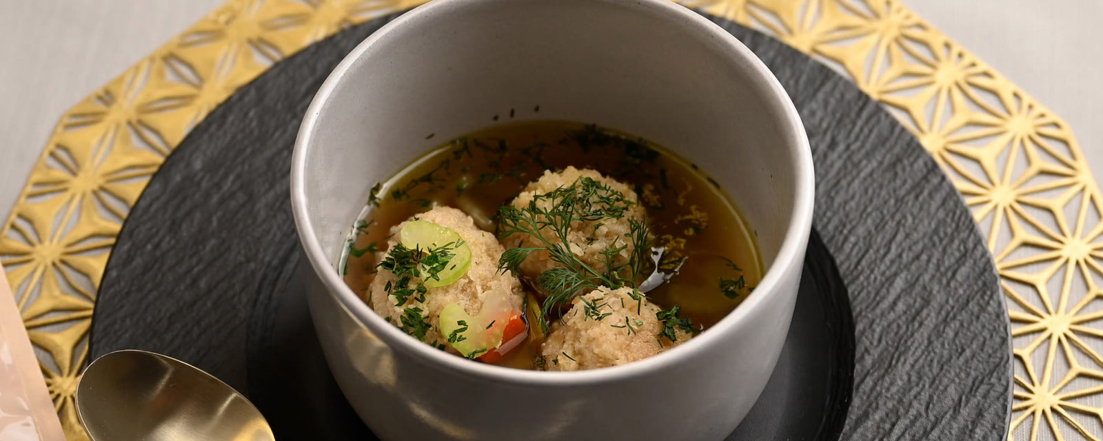 Herb-garnished Quinoa Matzoh Ball soup served in a modern gray cup with clear broth and vegetables.