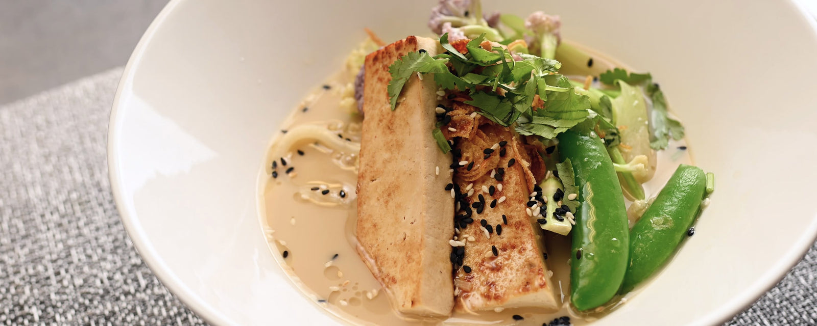 Soup noodle with tofu, snap peas, cilantro, sesame seeds, and crispy onions in a white bowl.