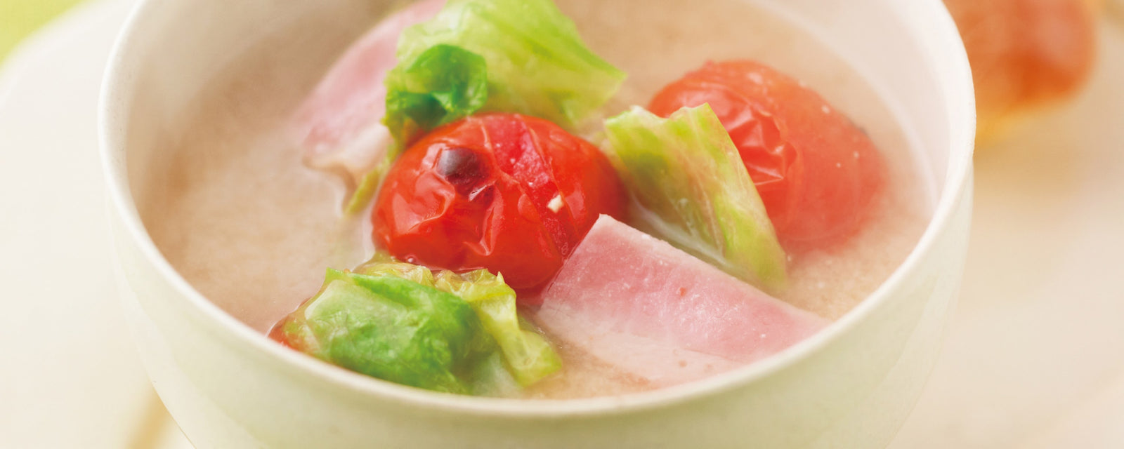 Miso soup with cherry tomatoes, lettuce pieces, and slices of bacon in a white bowl.