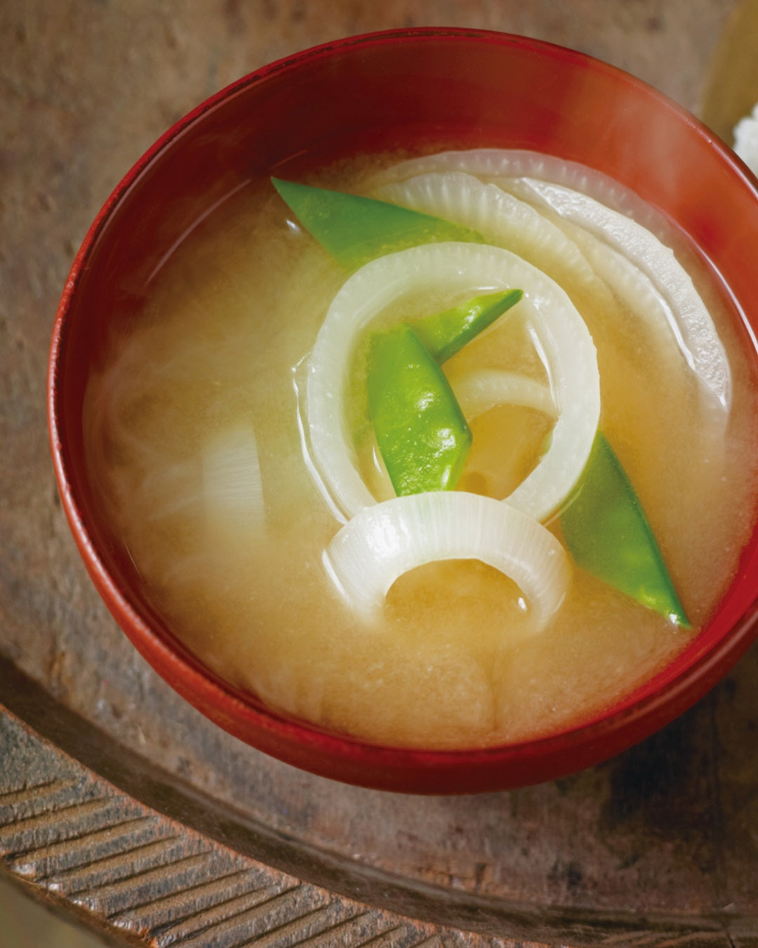 Miso soup with sliced onions and snap peas, served in a red bowl alongside rice balls.