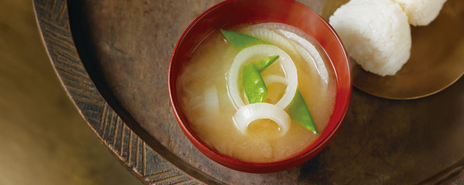 Miso soup with sliced onions and snap peas, served in a red bowl alongside rice balls.