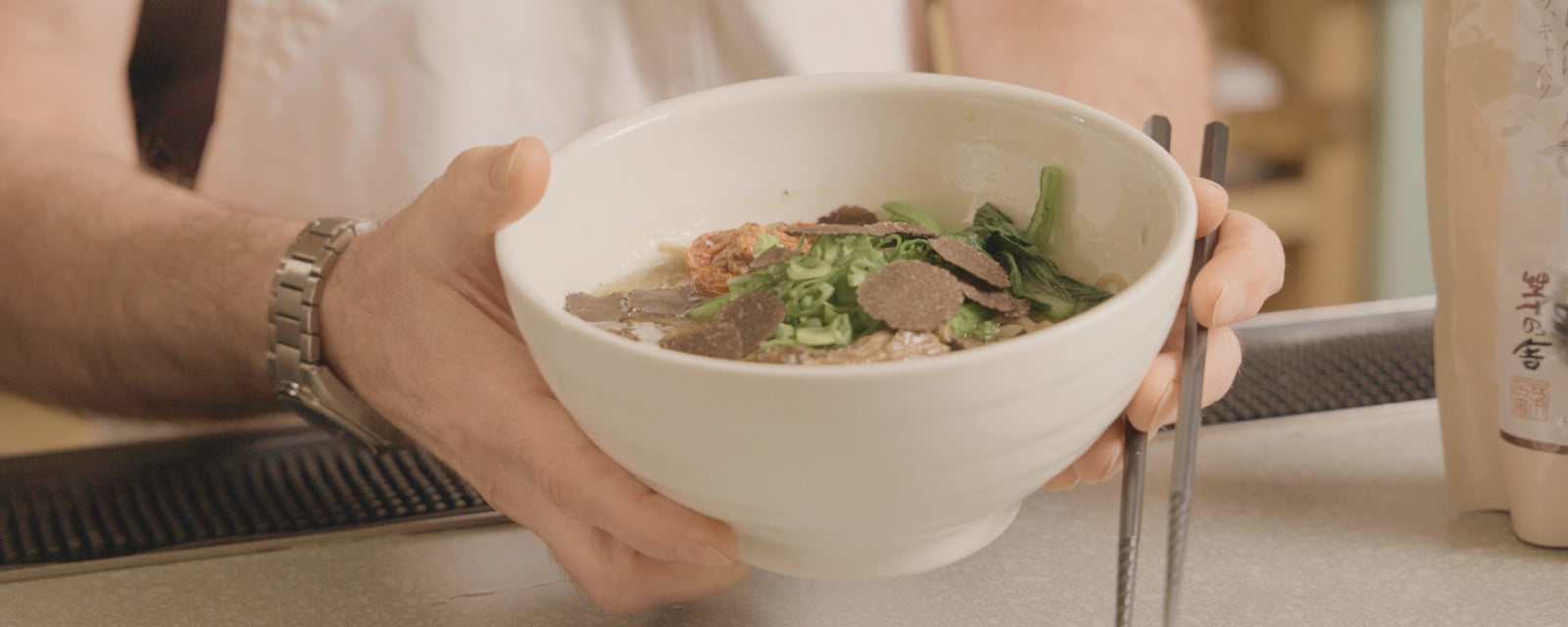 A person holding a bowl of truffle ramen topped with sliced truffle and green onions.