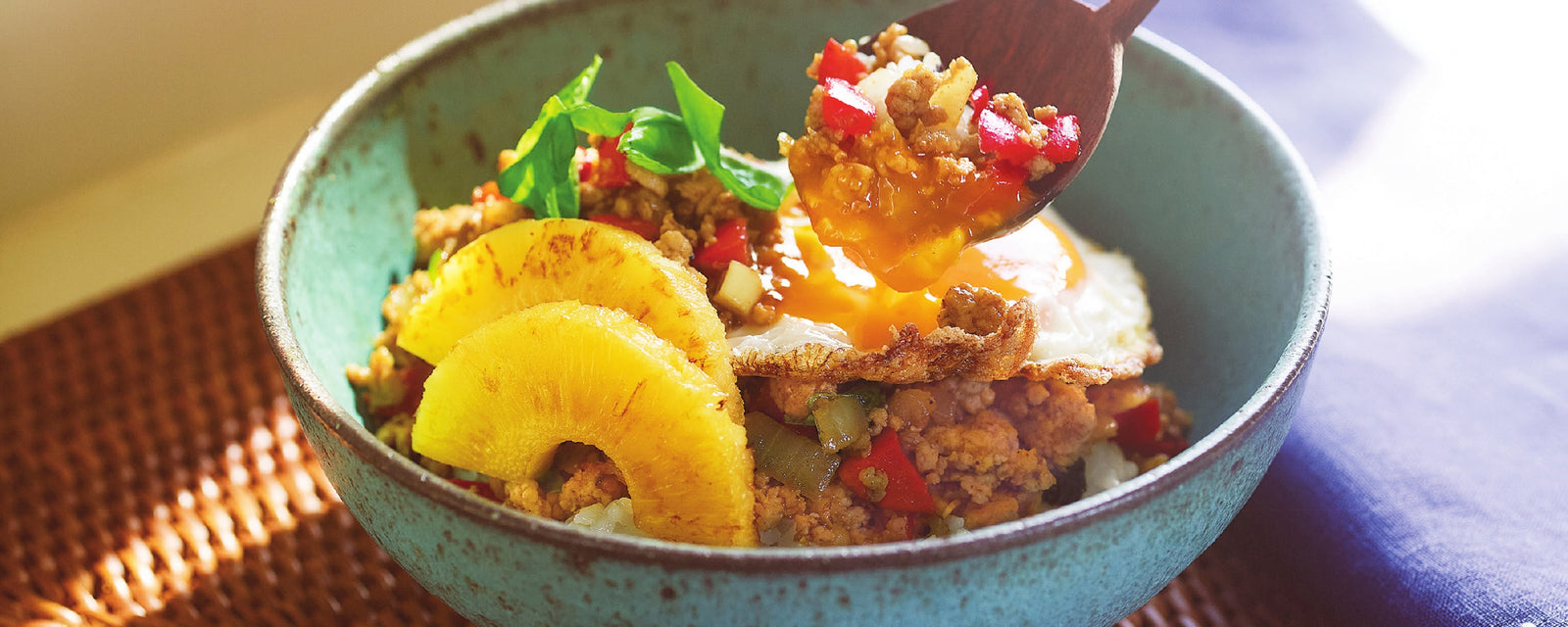 A bowl of Thai-inspired basil chicken rice topped with pineapple slices and a fried egg.