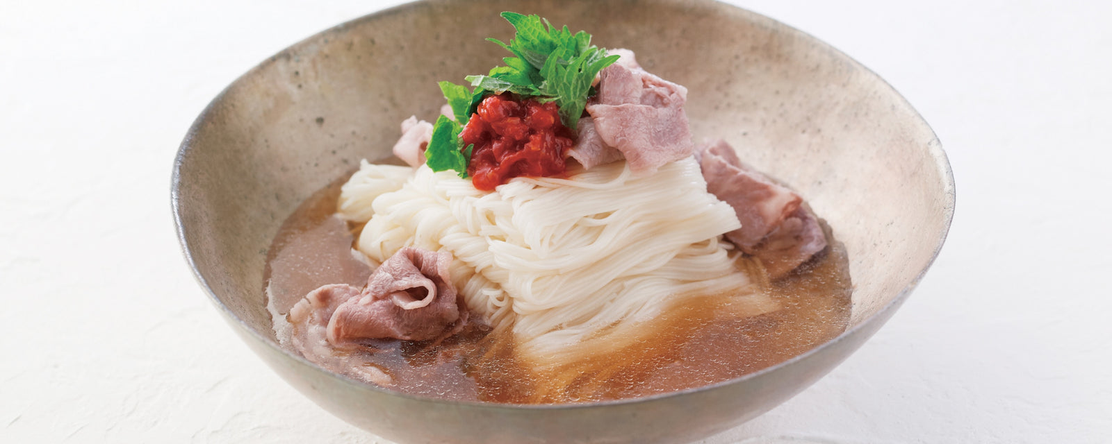A bowl of ume-spicy cold somen noodles topped with pork slices and fresh herbs in chilled broth.