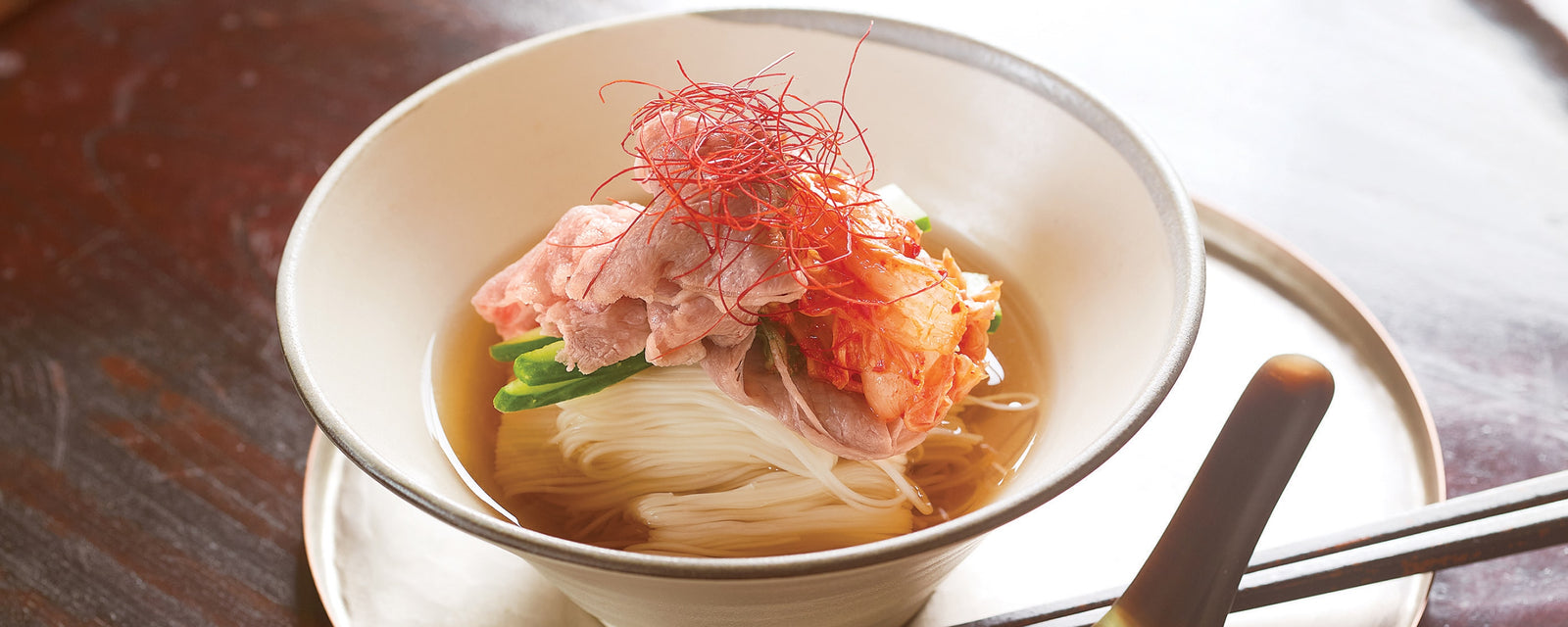 A bowl of Korean-style cold somen noodles topped with pork, kimchi, and chili threads in chilled broth.