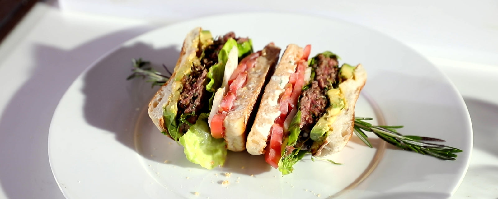 Beef hamburger sandwich with lettuce, tomato, and avocado, cut into pieces and served on a plate.