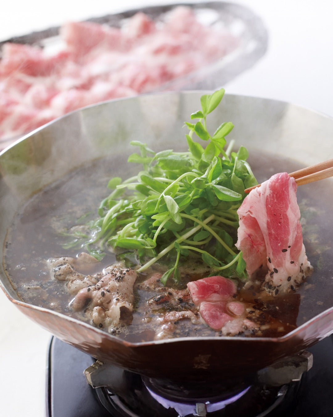Thinly sliced pork cooking in a black hot pot with greens, lifted with chopsticks.