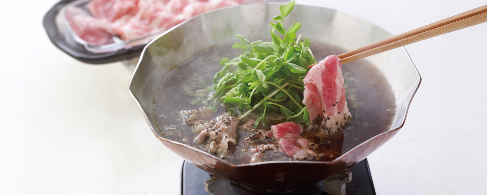 Thinly sliced pork cooking in a black hot pot with greens, lifted with chopsticks.