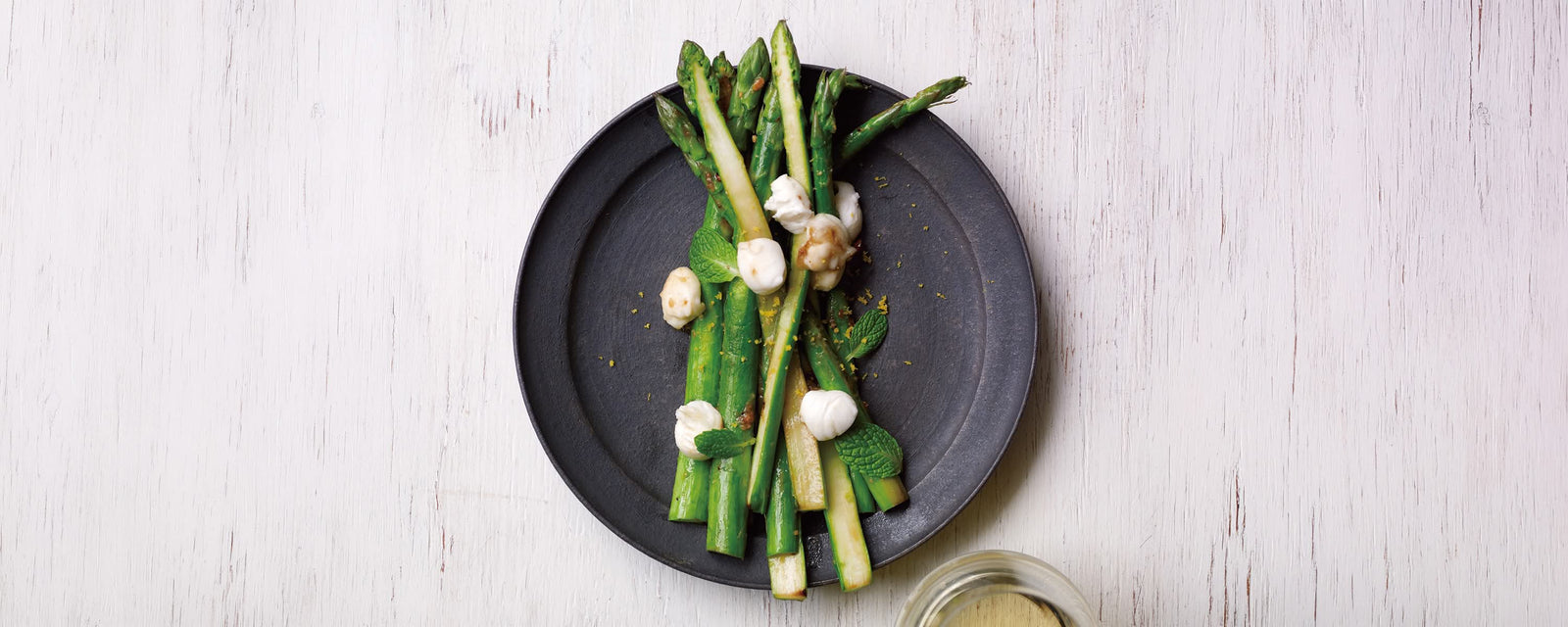 Sautéed green asparagus spears topped with melted mozzarella cheese, fresh mint leaves, and lemon zest on a white plate.