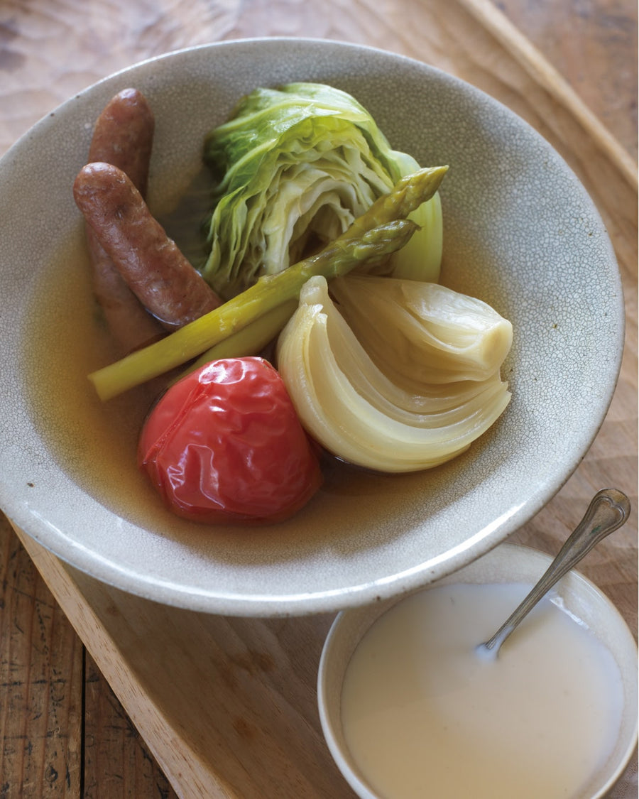 A steaming bowl of Japanese-style spring vegetable pot-au-feu featuring cabbage, carrots, potatoes, and sausages in a clear broth, served with a side of creamy garlic aioli sauce.