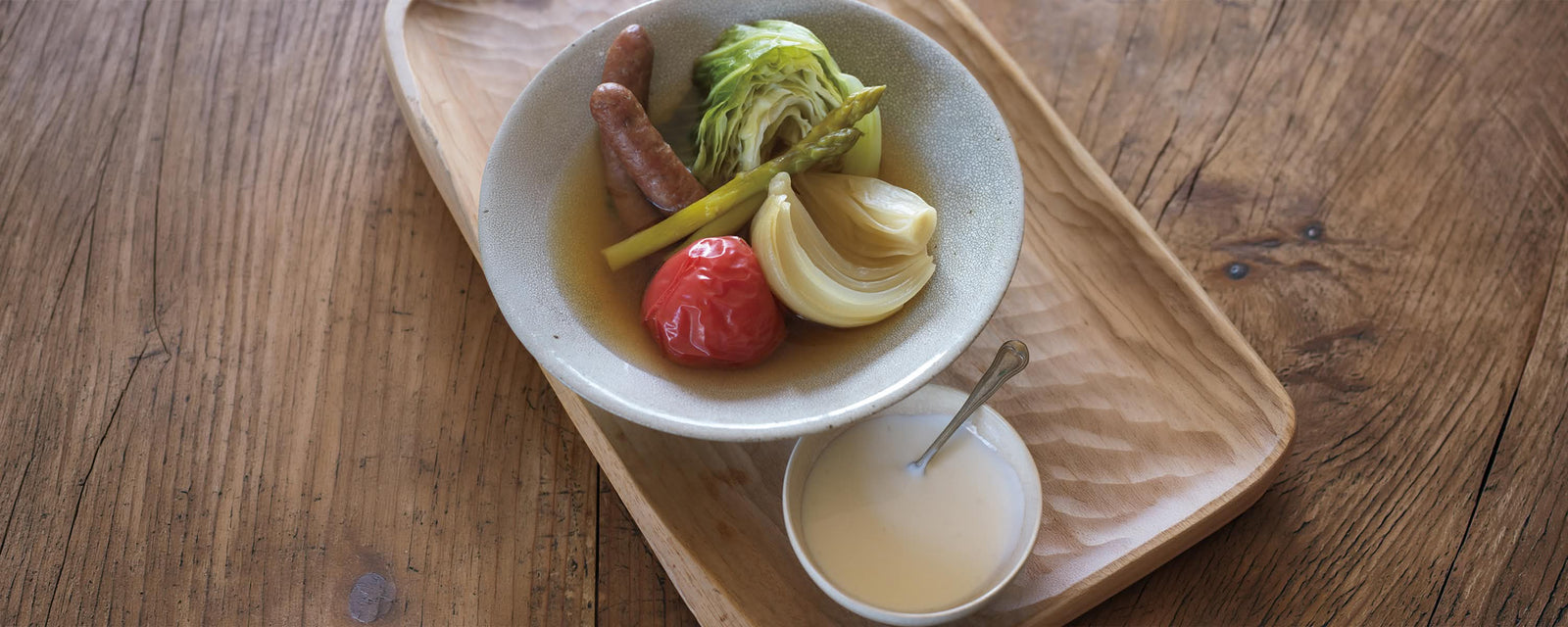 A steaming bowl of Japanese-style spring vegetable pot-au-feu featuring cabbage, carrots, potatoes, and sausages in a clear broth, served with a side of creamy garlic aioli sauce.