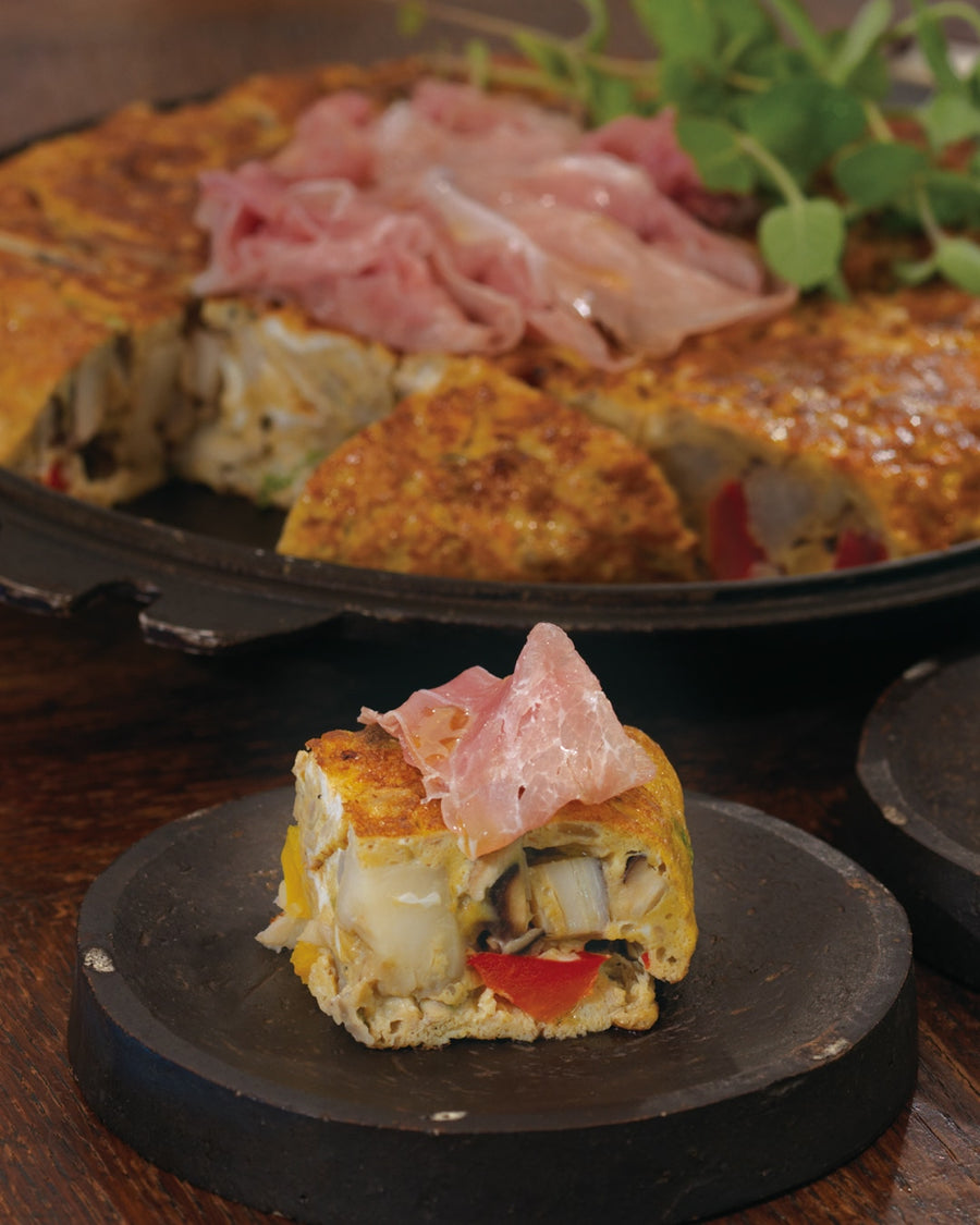 A colorful, large omelet filled with assorted vegetables, topped with thin slices of prosciutto and fresh herbs.