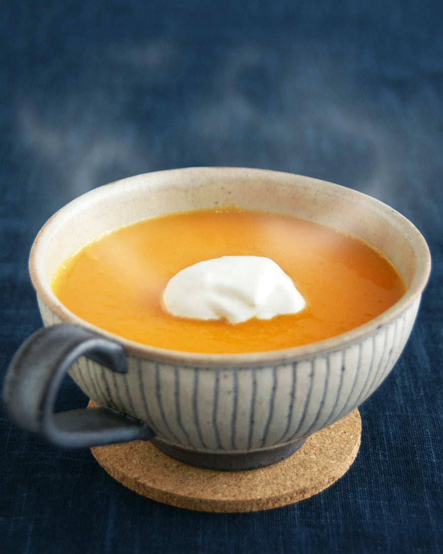 A cup of smooth, vibrant orange carrot soup topped with a dollop of cream, served in a striped ceramic bowl on a cork coaster against a dark blue background.