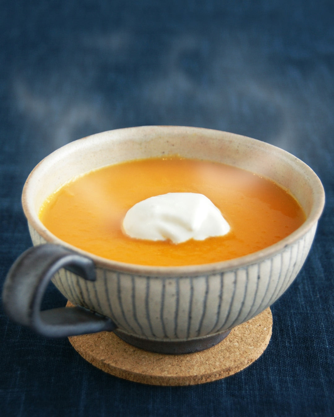 A cup of smooth, vibrant orange carrot soup topped with a dollop of cream, served in a striped ceramic bowl on a cork coaster against a dark blue background.