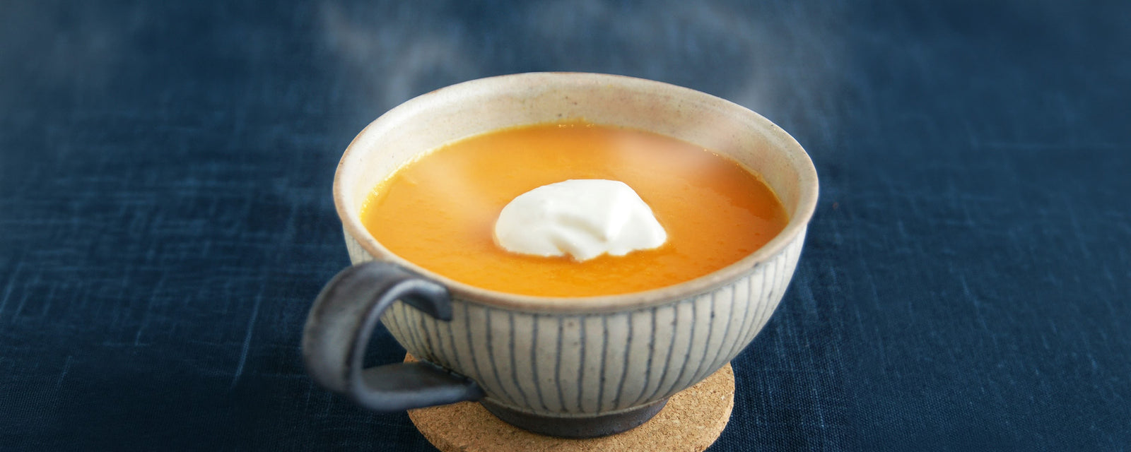 A cup of smooth, vibrant orange carrot soup topped with a dollop of cream, served in a striped ceramic bowl on a cork coaster against a dark blue background.