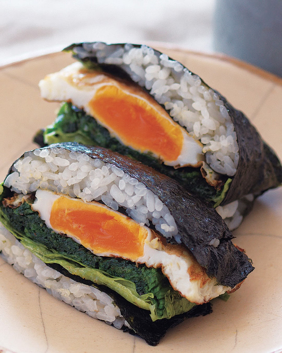 Two halves of a sushi sandwich wrapped in seaweed, filled with white rice, leafy greens, and a soft-boiled egg with a runny yolk, served on a beige plate.