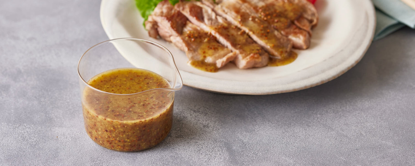 Sweet mustard dressing with whole grain mustard in a glass container, served with sliced pork sauté in the background
