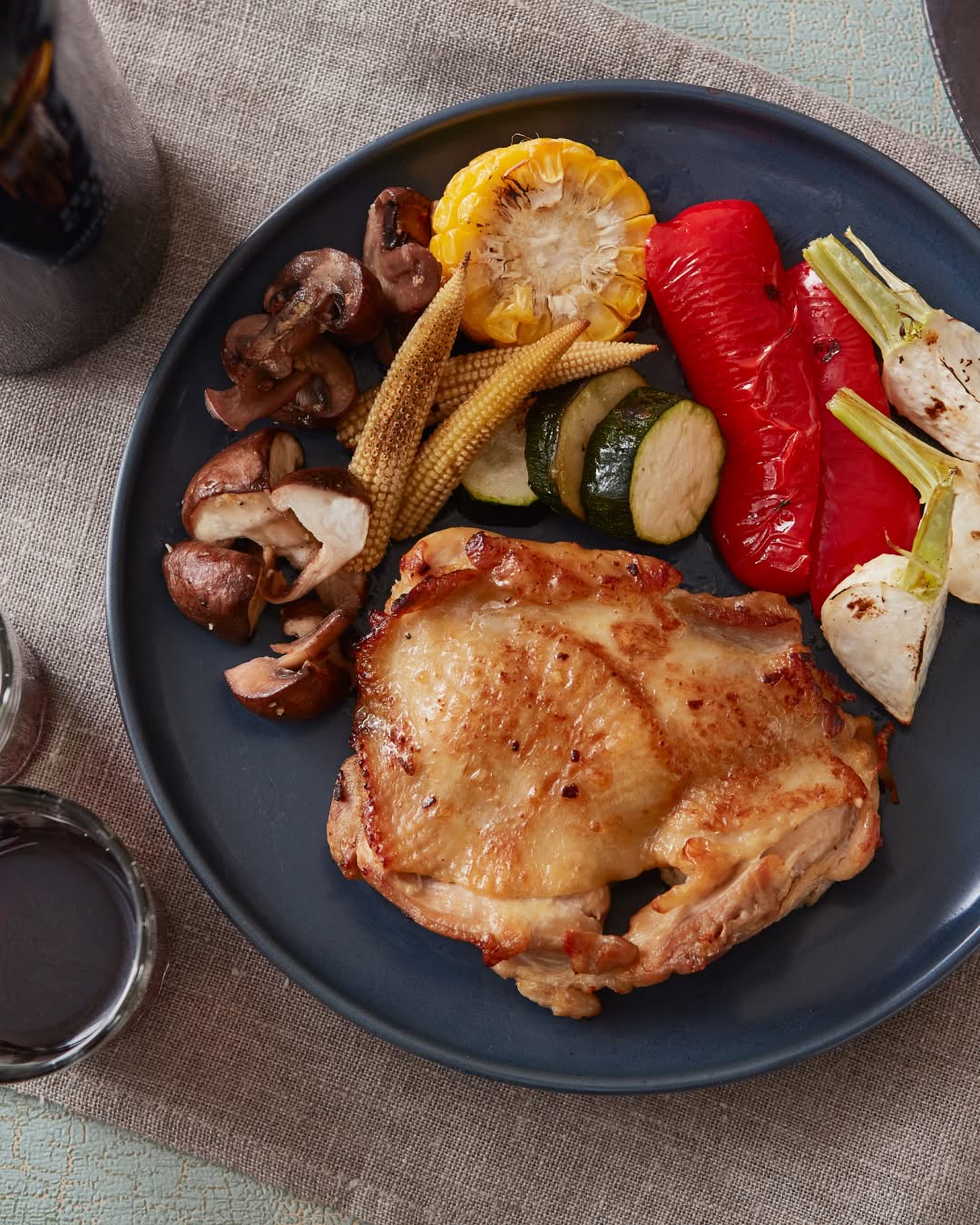 Roasted chicken thigh served with vegetables including mushrooms, baby corn, zucchini, and red pepper on a dark plate