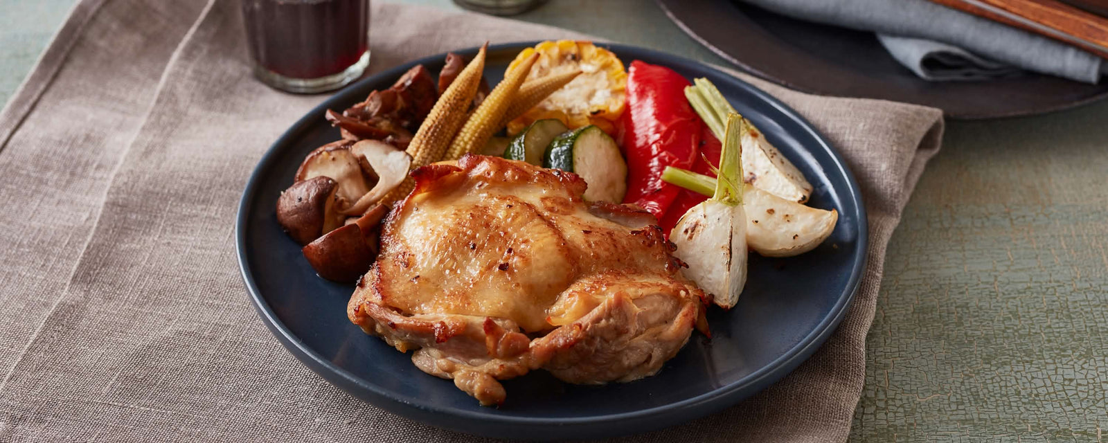 Roasted chicken thigh served with vegetables including mushrooms, baby corn, zucchini, and red pepper on a dark plate