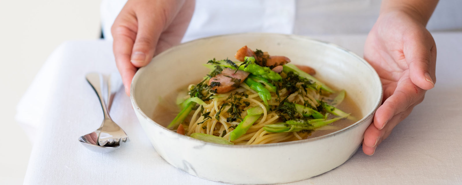 Hands holding a bowl of spaghetti in light umami dashi broth with asparagus, bacon, and shredded nori