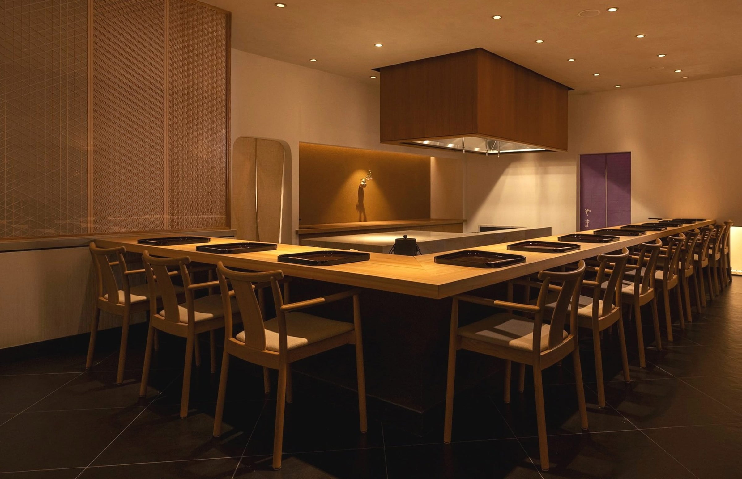 A modern, minimalist sushi restaurant interior with a U-shaped wooden counter, wooden chairs, black placemats, soft overhead lighting, and clean, neutral-toned walls creating an elegant and inviting dining space.