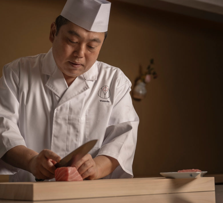 Chef in a traditional white uniform and hat skillfully cutting raw fish on a wooden board.