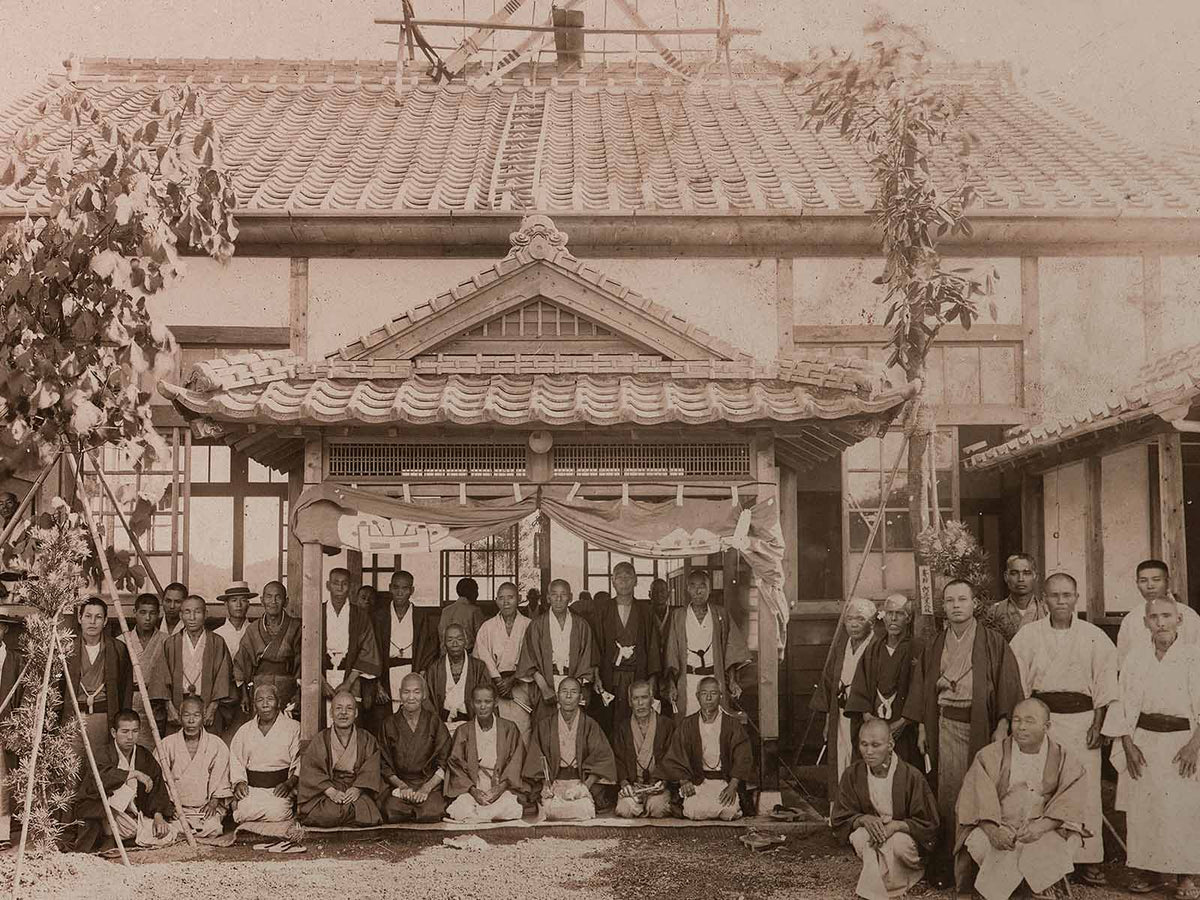 A sepia-toned historical photo showing a large group of men in traditional Japanese clothing posing in front of a wooden building with a tiled roof. Some men are seated while others stand behind them.