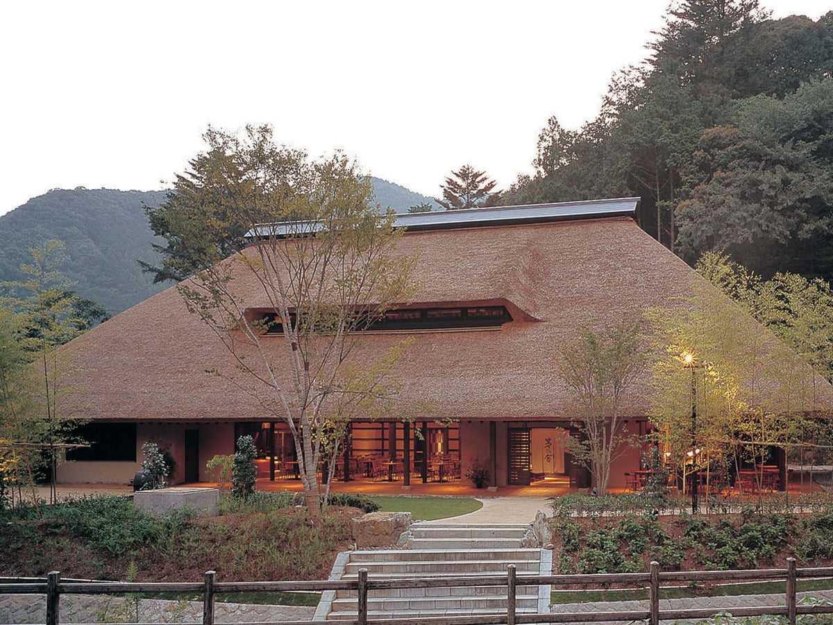 A large, traditional Japanese building with a thatched roof, warm lighting, and open entrance, surrounded by trees and mountains in the background.