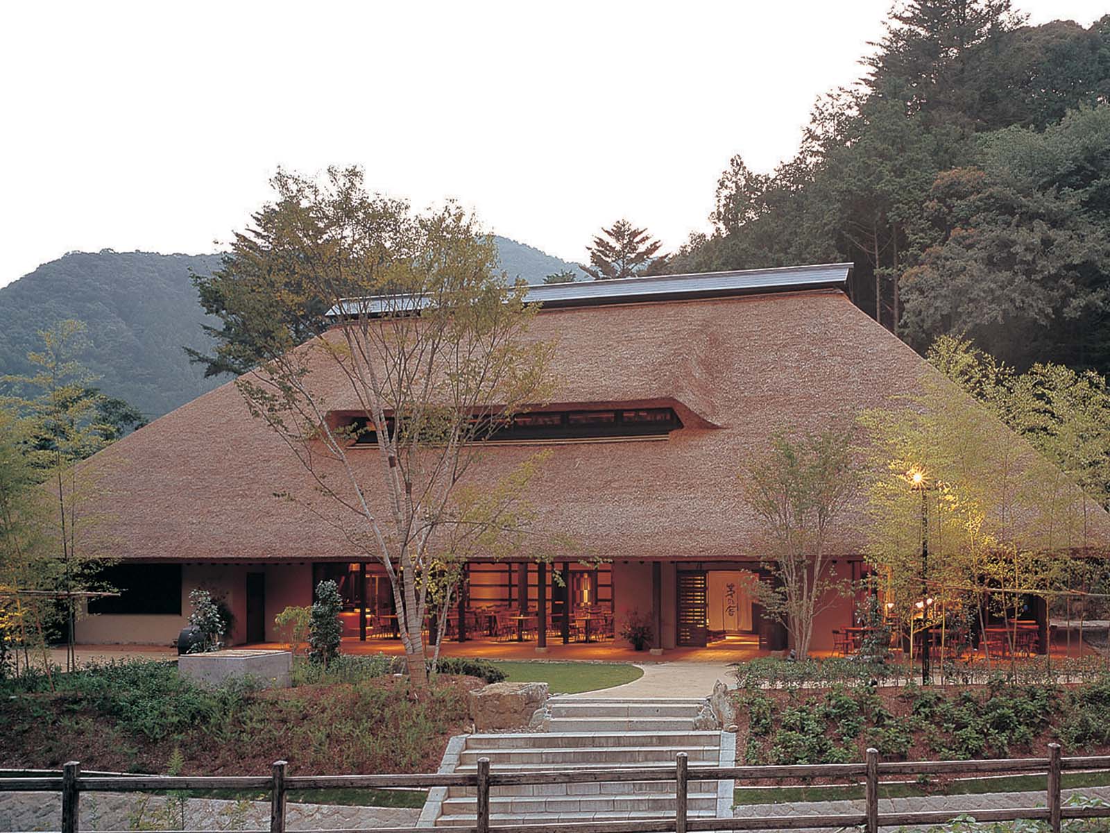A large, traditional Japanese building with a thatched roof, warm lighting, and open entrance, surrounded by trees and mountains in the background.