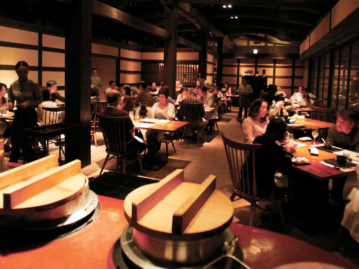 A warmly lit, busy restaurant with wooden beams and tables, where many people are dining and socializing. Traditional Japanese decor is visible, and food is being served to guests seated in groups.